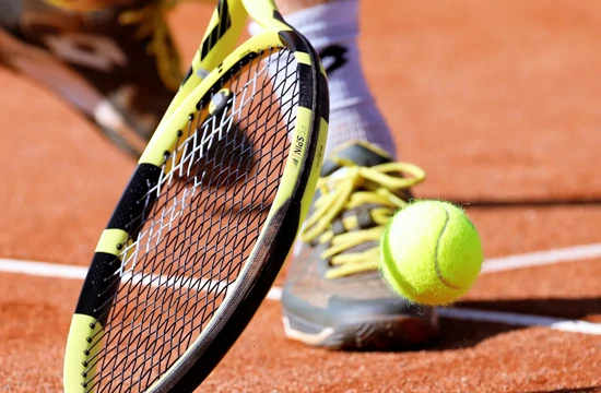 Les activités et loisirs autour de Lacanau. Deux pieds d'un joueur de tennis sur un terrain de terre battue, il reprend en demie volée une balle de tennis jaune avec une raquette noire et jaune.