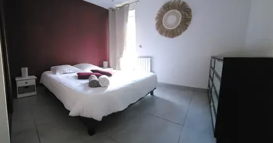 appart hôtel pasino n°7, chambre avec grand lit double 160/200 donnant sur la patio. Décoration dans les tons bordeaux avec 2 coussins sur le lit et 2 serviettes de bain blanche et grise. Chambre entièrement carrelée au sol (couleur grise). Sur la droite une commode 8 tiroirs. Un rideau occultant beige est posé devant la porte vitrée donnant sur le patio