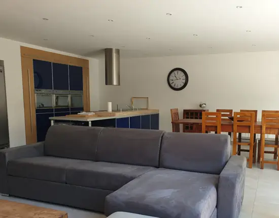 appart hôtel pasino n°7, à louer pour vos vacances. Ici vous pouvez voir la grande pièce à vivre comportant une grande cuisine ouverte toute équipée avec un grand îlot dans les ton bois et bleu, un espace séjour avec une grande table en bois et ses chaises en bois, ainsi qu'un grand canapé d'angle en velours gris anthracite