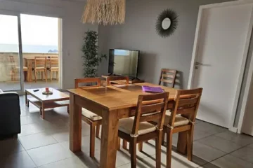 Appart hôtel Pasino N°2, pièce à vivre de l'appartement numéro 2, elle est lumineuse avec ses murs blancs,. La pièce est meublée avec des meubles en teck: une table, ses six chaises et leurs coussin d'assise beiges. A l'arrière la partie salon : sa table de salon en teck , son meuble TV et la TV grand écran. Des éléments de décoration au mur et au plafond, un palmier au fond de la pièce. une baie vitrée donne sur la terrasse vue mer et ses meubles en teck