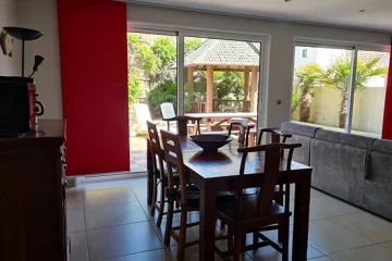 appart hôtel pasino n°8, pièce à vivre. Elle donne sur un patio ensoleillé via une grande baie vitrée protégée du soleil par des rideaux japonais rouges. les meubles sont en teck. Une joli table de repas et ses six chaises. Au fond à droite, un grand canapé et sa méridienne sépare la partie repas et la partie salon