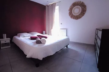 appart hôtel pasino n°7, chambre avec grand lit double 160/200 donnant sur la patio. Décoration dans les tons bordeaux avec 2 coussins sur le lit et 2 serviettes de bain blanche et grise. Chambre entièrement carrelée au sol (couleur grise). Sur la droite une commode 8 tiroirs. Un rideau occultant beige est posé devant la porte vitrée donnant sur le patio