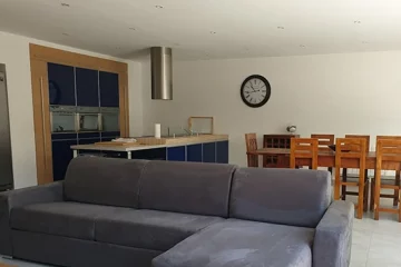 appart hôtel pasino n°7, pièce principale de l'appartement n°7. Au premier plan le canapé et sa méridienne gris. A l'arrière la grande table et ses 8 chaises en Teck sur la gauche la grande cuisine équipée Laquée bleu. Au mur une grande pendule