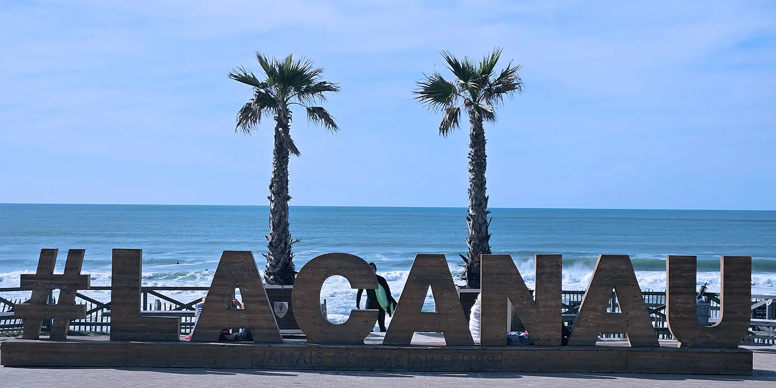 Lacanau , jamais comme ailleurs, sculpture en bois situées sur le front de mer au dessus de la plage centrale 2 palmiers derrière la structure, l'océan avec des couleurs qui vont du turquoise au bleu profond, un ciel bleu lumineux.