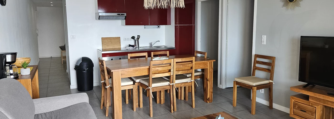 appart hôtel pasino n°3, pièce principale, au premier plan le coin salon et ses meuble en teck, son palmier et sa TV grand écran. A gauche une tables et ses chaises et la Kitchenette équipée aux façades rouges. A gauche une console en teck avec des éléments de décoration