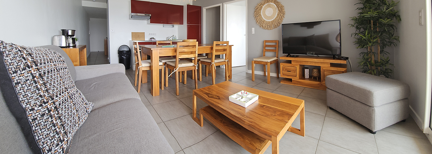 appart hôtel pasino n°3, pièce principale, au premier plan le coin salon et ses meuble en teck, son palmier et sa TV grand écran. A gauche une tables et ses chaises et la Kitchenette équipée aux façades rouges. A droite une console en teck avec des éléments de décoration