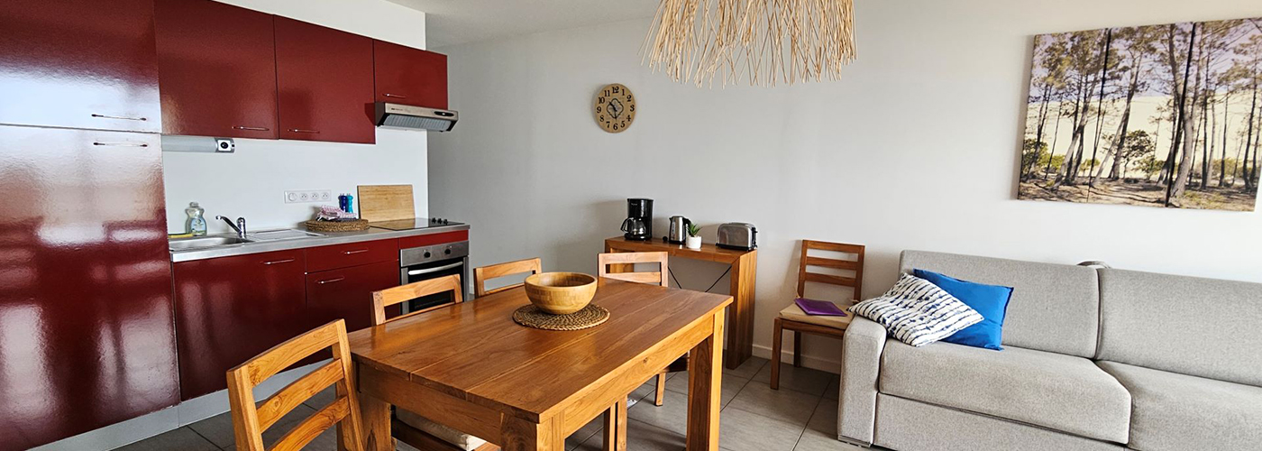 Appart hôtel Pasino n°5 pièce à vivre A gauche, la kitchenette équipée rouge, au milieu une table en teck et ses chaise. Sur la droite à l'avant un canapé convertible; Sur le mur une des éléments de décoration