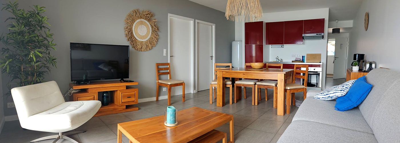 Appart hôtel Pasino n°5 pièce à vivre Au fond, la kitchenette équipée rouge, au milieu une table en teck et ses chaise. Sur la gauche à l'avant un fauteuil tournant blanc, une table basse en teck posé dessus un élément de décoration en verre bleu. Le long du mur au fond,, un palmier, un meuble TV et sa TV grand écran. Sur le mur une décoration exotique
