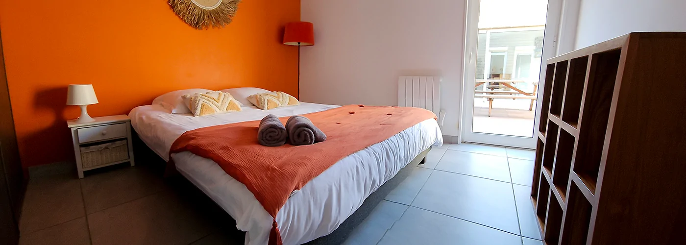 appart hôtel Pasino n°7, chambre avec grand lit double 160/200 donnant sur la patio. Décoration dans les tons orangés avec plaid orange sur le lit et 2 serviettes de bain grise. Chambre entièrement carrelée au sol (couleur grise). Un meuble de rangement en tecks