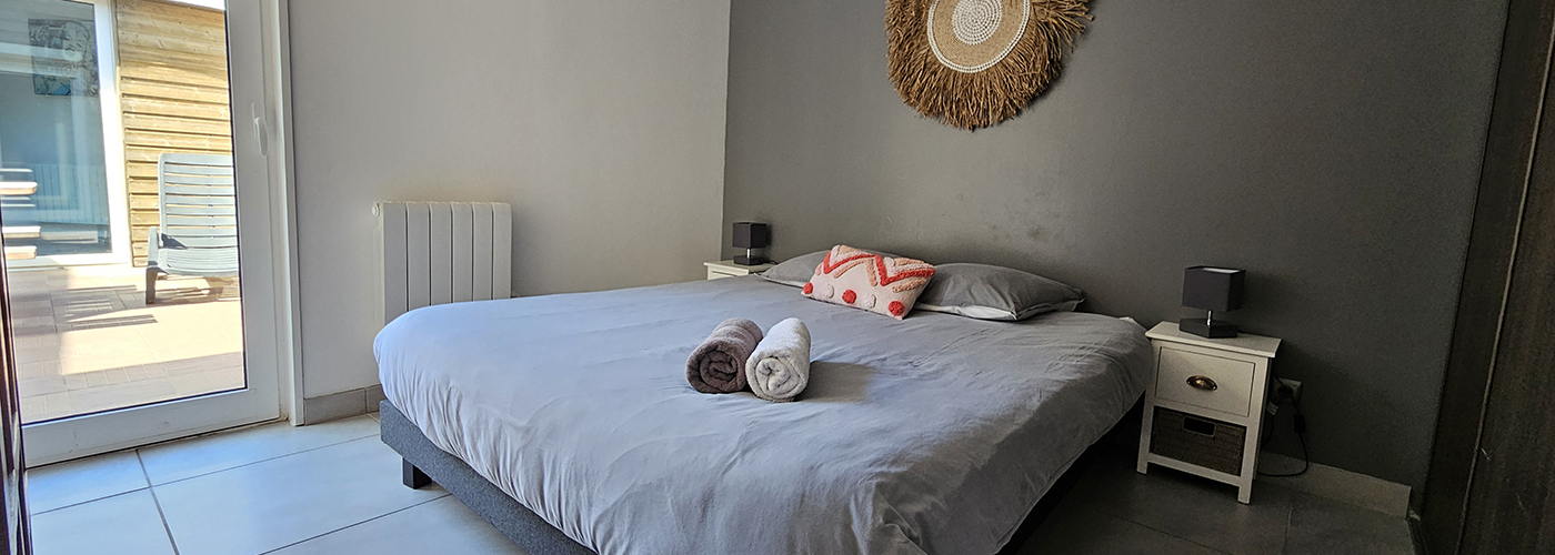 appart hôtel pasino n°7, chambre avec grand lit double 160/200 donnant sur la patio. Décoration dans les tons gris avec un coussin de décoration et 2 serviettes de bain, une grise et une blanche. de chaque coté du lit un chevet 2 tiroir et sa lampe. Chambre entièrement carrelée au sol (couleur grise) Au mur une décoration exotique.