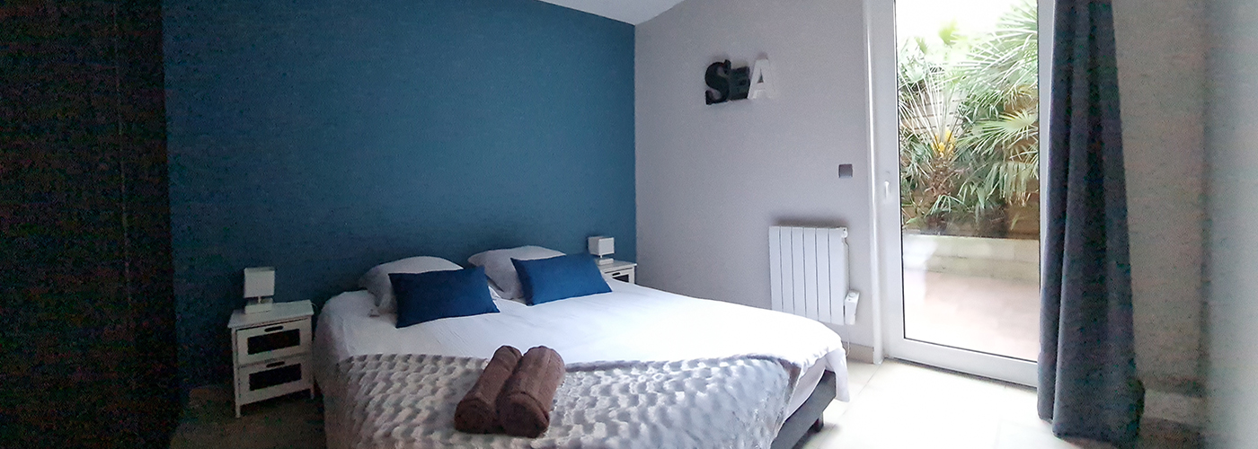 appart hôtel pasino n°8, chambre aux tons dominante de bleus, au mur une décoration "sea". La chambre est composée d'un grand lit et ses 2 chevets blancs à tiroirs gris et leur lampe. Sur le lit, 2 coussins de décoration bleuet, un plaid gris et ses 2 serviettes grises posées dessus. A l'arrière une porte fenêtre et son rideau occultant bleu un patio et ses palmiers