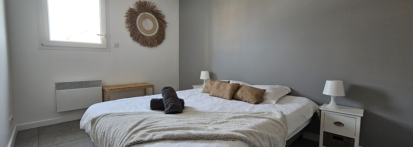 appart hôtel pasino n°3, chambre N°3 murs blancs et gris, une décoration exotique sur le mur blanc dessous un banc en pin, une suspension au plafond, 2 chevet plus lampes, un lit en 160X200 , 2 oreillers, 3 coussins beiges, un plaid et 2 serviettes sur le lit