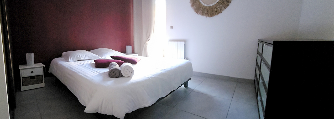 appart hôtel pasino n°7, chambre avec grand lit double 160/200 donnant sur la patio. Décoration dans les tons bordeaux avec 2 coussins sur le lit et 2 serviettes de bain blanche et grise. Chambre entièrement carrelée au sol (couleur grise). Sur la droite une commode 8 tiroirs. Un rideau occultant beige est posé devant la porte vitrée donnant sur le patio