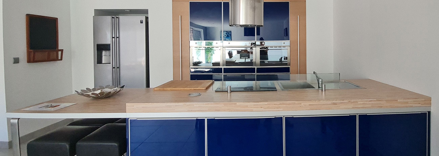 appart hôtel pasino n°7, la grande cuisine aux façades bleues laquées. De nombreux placards de rangement. Un plan de travail couleur bois. sur l'ilot central. Un évier et la table de cuisson sont au centre de celui-ci. au dessus une hotte en inox. A l'arrière, un rectangle de placard couleur bois entoure un ensemble de placard, en haut de fours au centre et à nouveau placard de rangement en bas ainsi que le lave-vaisselle. A gauche le réfrigérateur américain.