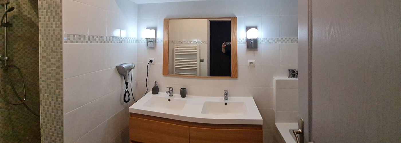 appart hôtel pasino n°7, salle de bain, au premier plan, une double vasque, en face, un miroir entouré de bois beige de chaque coté du miroir des appliques argentées avec un globe blanc., juste en dessous une prise électrique. On voit dans le reflet du miroir un radiateur électrique. Sur la paroi du mur, un sèche-cheveux. A gauche la douche italienne et à droite la baignoire. Sous les double vasques, un meuble sous vasque 4 tiroirs en bois clair. Une poubelle est posée au sol.
