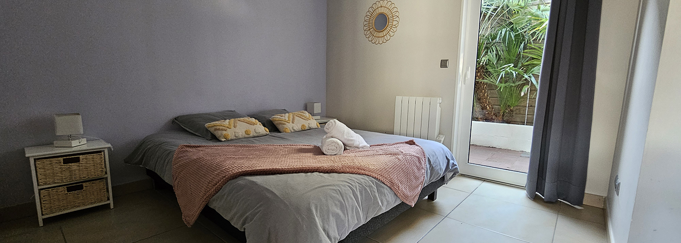 appart hôtel pasino n°8, chambre aux tons dominante de gris, au mur une décoration miroir soleil entouré de rotin. La chambre est composée d'un grand lit et ses 2 chevets blancs à tiroirs en rotin et leur lampe. Sur le lit , 2 coussins de décoration déco ethnique , 2 serviettes blanches posées sur un plaid ROSE. A l'arrière une porte fenêtre et son rideau occultant gris donnant sur un patio et ses palmiers