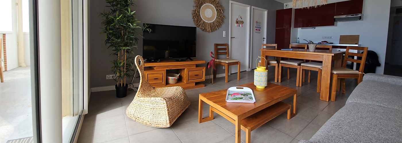 Appart hôtel Pasino n°4 Pièce à vivre, elle est lumineuse avec ses murs blancs, elle est meublée avec des meubles en teck: une table, ses six chaises et leurs coussins d'assise beiges. A l'avant, la partie salon: sa table de salon en teck sur la table un plateau coloré et une lanterne jaune , son meuble TV et la TV grand écran. des éléments de décoration au mur, un palmier et un fauteuil en rotin à gauche de la pièce., à droite assise du canapé gris sur lequel est posé un coussin jaune. Au fond, vue sur l'entrée de l'appartement et la kitchenette rouge.