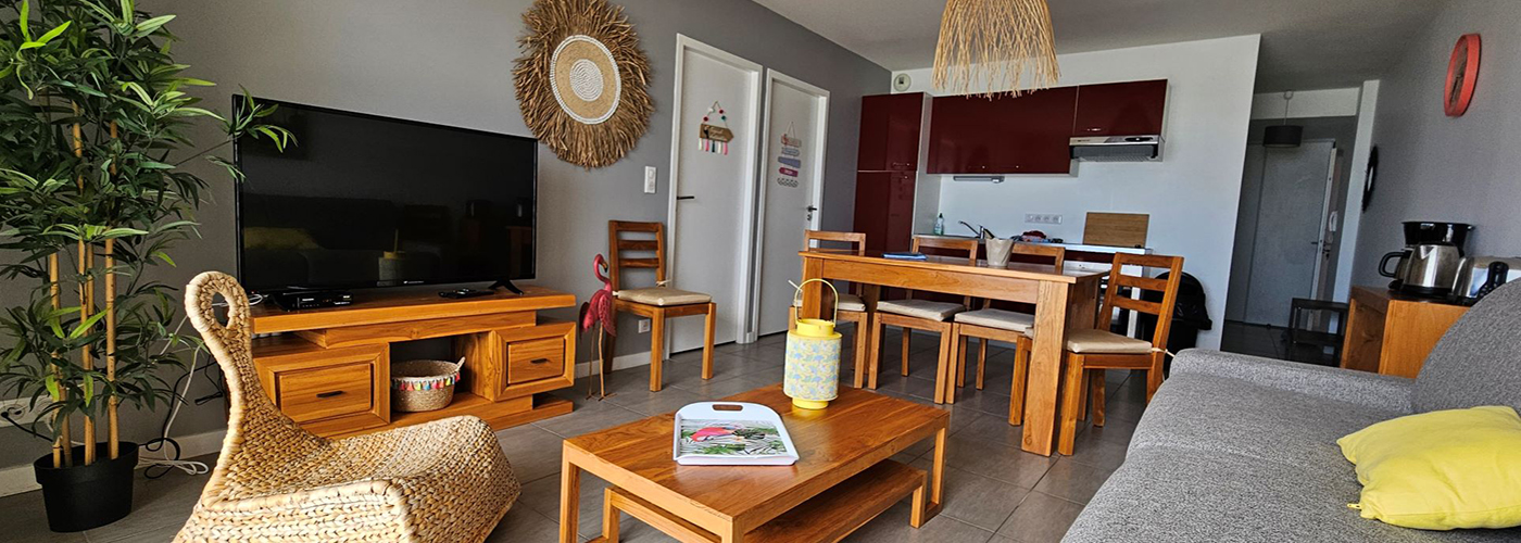 Appart hôtel Pasino n°4 Pièce à vivre, elle est lumineuse avec ses murs blancs, elle est meublée avec des meubles en teck: une table, ses six chaises et leurs coussins d'assise beiges. A l'avant, la partie salon: sa table de salon en teck sur la table un plateau coloré et une lanterne jaune , son meuble TV et la TV grand écran. des éléments de décoration au mur, un palmier et un fauteuil en rotin à gauche de la pièce., à droite assise du canapé gris sur lequel est posé un coussin jaune. Au fond, vue sur l'entrée de l'appartement et la kitchenette rouge.