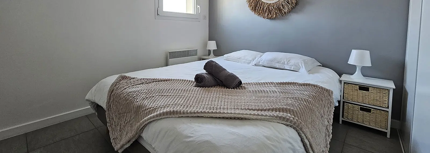 appart hôtel pasino n°6, chambre N°2 murs blancs et gris, une décoration exotique sur le mur blanc, une suspension au plafond, 2 chevets blancs : 2 tiroirs en Osiers plus lampes blanches, un lit en 160X200 , 2 oreillers, un plaid beige et 2 serviettes sur le lit