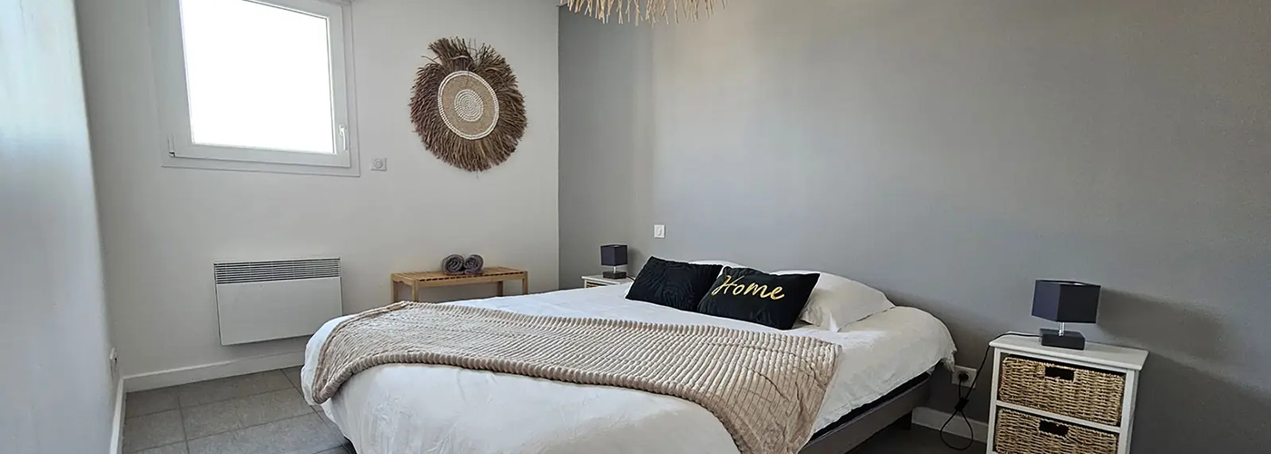 appart hôtel pasino n°6, chambre N°3 murs blancs et gris, une décoration exotique sur le mur blanc dessous un banc en pin, une suspension au plafond, 2 chevets blancs : 2 tiroirs en Osiers plus lampes grises, un lit en 160X200 , 2 oreillers, 2 coussins décoration Vert foncé l'un marqué "Home", 2 serviettes sur le lit