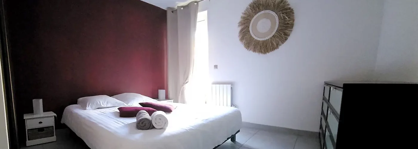 Appart-hotel-appartement-n7-chambre1 appart hôtel pasino n°7, chambre avec grand lit double 160/200 donnant sur la patio. Décoration dans les tons bordeaux avec 2 coussins sur le lit et 2 serviettes de bain blanche et grise. Chambre entièrement carrelée au sol (couleur grise). Sur la droite une commode 8 tiroirs. Un rideau occultant beige est posé devant la porte vitrée donnant sur le patio