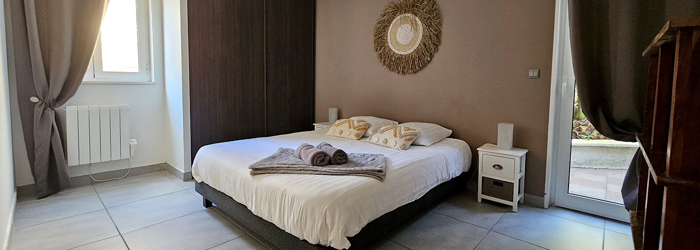 appart hôtel pasino n°7, appartement N°7 Chambre aux tons dominante de beige foncé, au mur une décoration miroir soleil entouré de rotin. La chambre est composée d'un grand lit et ses 2 chevets blancs à tiroirs en rotin et leur lampe. Sur le lit 2 coussins de décoration déco ethnique , 2 serviettes grises posées sur un plaid gris. A l'arrière une porte fenêtre et son rideau occultant beige donnant sur un petit patio et ses palmiers