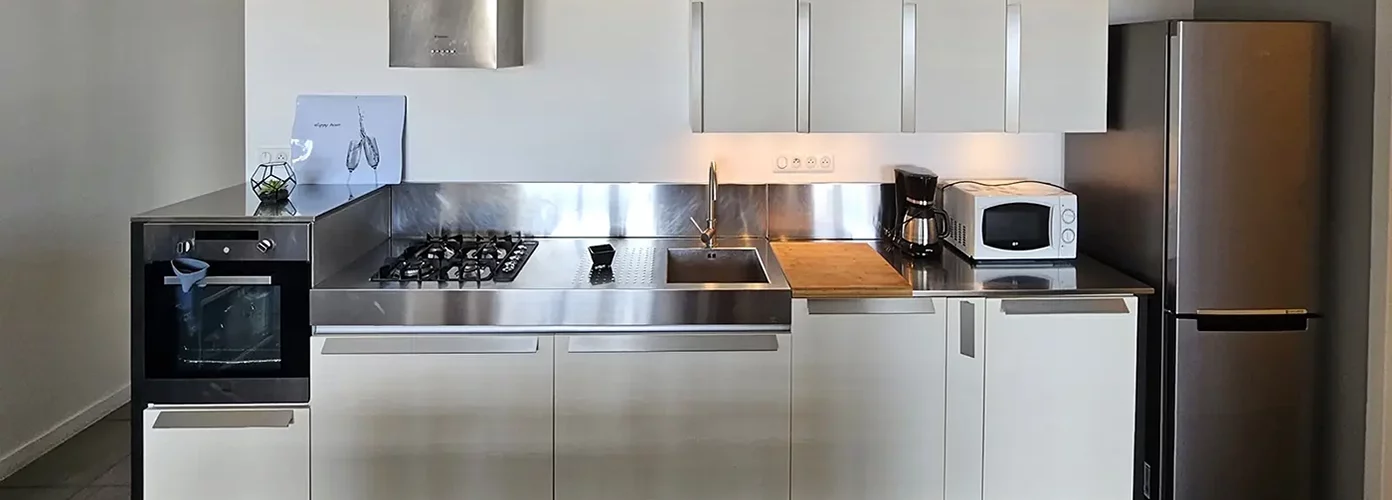 appart hôtel pasino n°6, cuisine de l'appartement sur un linéaire, à droite le réfrigérateur façade inox. les meubles de cuisine sont beiges. Sur le plan de travail en inox, un micro onde, une cafetière et une planche à découper en bois, un évier en inox, une table de cuisson au gaz, au dessus une hotte et un four