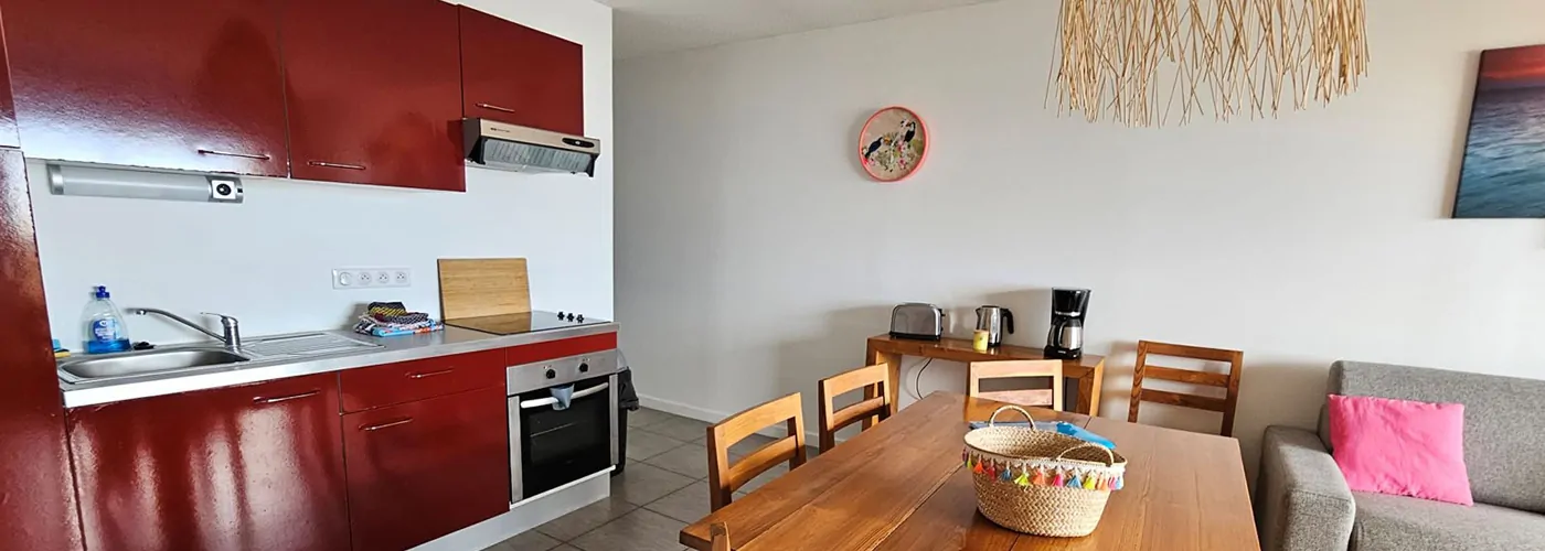 Appart hôtel Pasino n°4 pièce à vivre A gauche, la kitchenette équipée rouge, au milieu une table en teck et ses chaise. Sur la droite à l'avant un canapé convertible; Sur le mur une des éléments de décoration