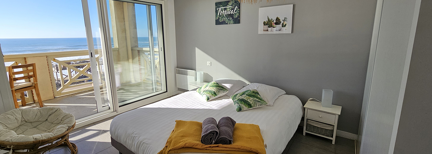 Appart Hôtel Pasino N°4, chambre appartement n°4 donnant sur la terrasse vue mer, 2 chevets, 2 lampes de chevet, 2 coussins décoration sur le lit, un plaid jaune, 2 serviettes posées sur le lit. Décorations au mur et plafond. sur la gauche un fauteuil en rotin. A l'arrière de la photo vue sur la terrasse et l'océan