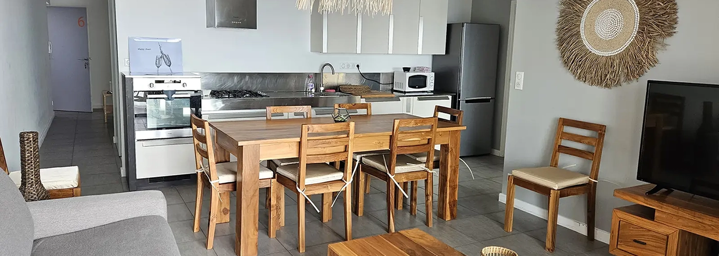 appart hôtel pasino n°6, pièce principale , un canapé gris, des meubles en teck, un écran TV, au milieu une table en teck et ses 8 chaises, au fond la cuisine équipée de meubles beiges et inox. Présence d'éléments de décoration aux murs et plafond.