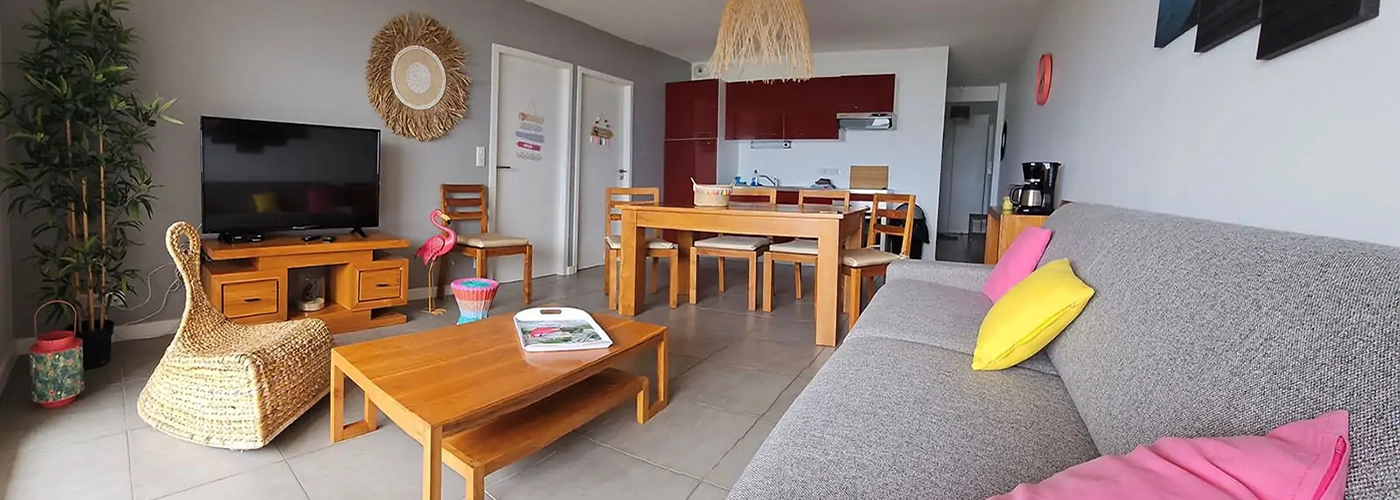 Appart hôtel Pasino n°1 Pièce à vivre, elle est lumineuse avec ses murs blancs, elle est meublée avec des meubles en teck: une table, ses six chaises et leurs coussin d'assise beiges. A l'avant, la partie salon: sa table de salon en teck sur la table un plateau coloré et une lanterne jaune , son meuble TV et la TV grand écran. des éléments de décoration au mur, un palmier et un fauteuil en rotin à gauche de la pièce., à droite assise du canapé gris sur lequel est posé un coussin jaune. Au fond, vue sur l'entrée de l'appartement et la kitchenette rouge.