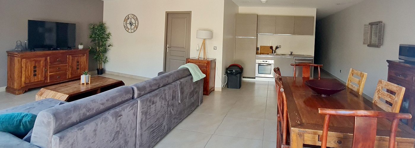 appart hôtel pasino n°8, pour vos vacances. Ici vous pouvez voir la pièce à vivre comprenant un espace séjour avec une table et ses chaises en bois, ainsi qu'un grand canapé d'angle en velours gris anthracite, ses meubles en teck et son grand écran TV. Des éléments de décorations agrémentent l'espace et le rendent chaleureux Au fond l'espace cuisine