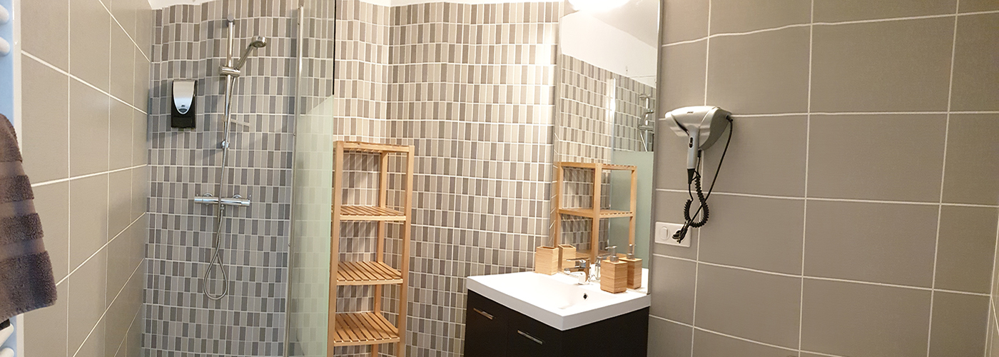 Appart hôtel Pasino n°1 Meuble vasque et son miroir, à sa gauche une étagère de rangement en bois et une douche italienne équipée d'une paroi anti projection en verre