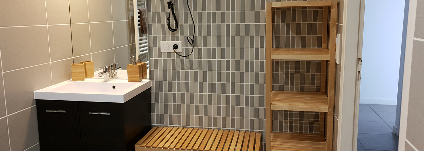 Appart hôtel Pasino n°6 Meuble vasque et son miroir, à sa droite, un sèche cheveux et des étagères de rangement en bois.