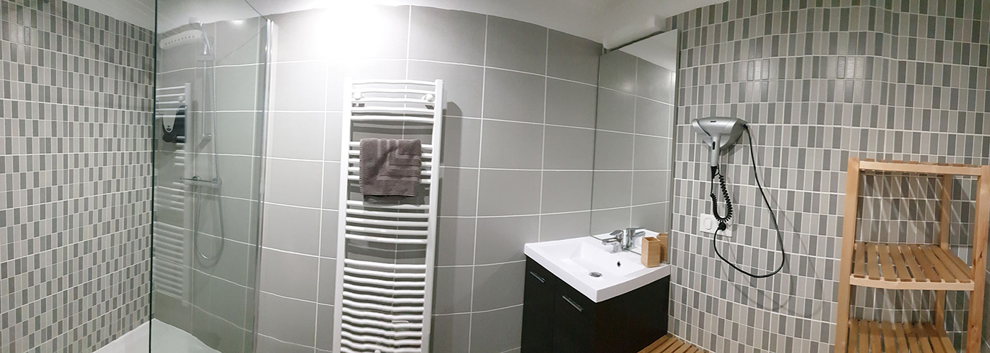 Appart hôtel Pasino n°3 Meuble vasque et son miroir, à sa droite, un sèche cheveux et des étagères de rangement en bois. A sa gauche un radiateur sèche serviettes et une douche italienne équipée d'une paroi anti projection en verre