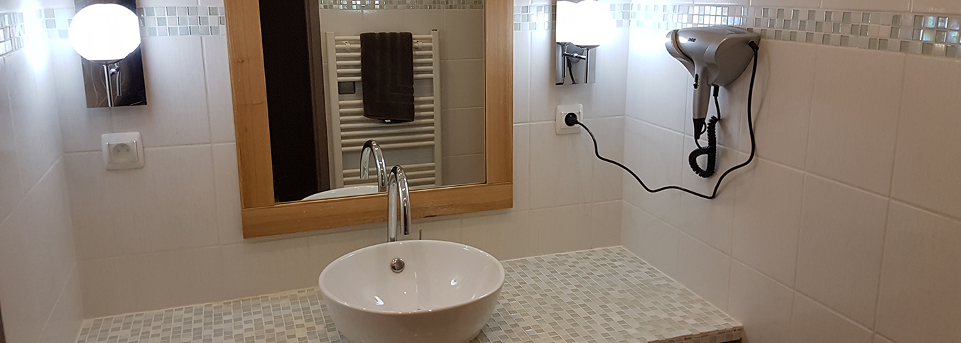 Salle de bain un mélange de petits et moyens carreaux blancs et verts très clair. Au centre un meuble et sa vasque au dessus un miroir, entouré de 2 appliques. Accroché au mur à droite, un sèche cheveu