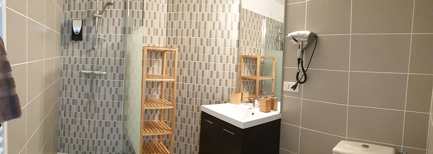 Appart hôtel Pasino n°1 Meuble vasque et son miroir, à sa gauche une étagère de rangement en bois et une douche italienne équipée d'une paroi anti projection en verre