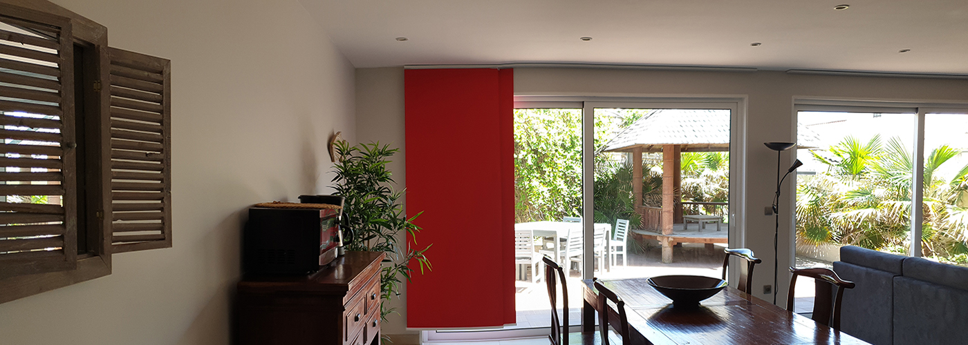appart hôtel pasino n°8, pièce à vivre de l'appartement. Elle donne sur un patio ensoleillé via une grande baie vitrée protégée du soleil par des rideaux japonais rouges. A gauche un palmier, tous les meubles sont en teck. Une joli table de repas et ses six chaises. Au fond à droite, un grand canapé et sa méridienne sépare la partie repas et la partie salon avec sa table basse, son grand meuble et son grand écran TV. Lampe et décorations moderne et exotique