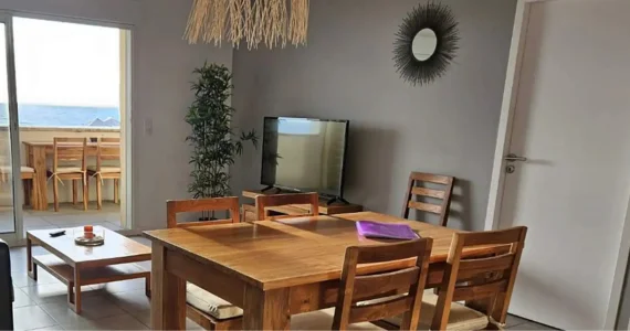 Appart hôtel Pasino n°2 Pièce à vivre, elle est lumineuse avec ses murs blancs. La pièce est meublée avec des meubles en teck: une table, ses six chaises et leurs coussins d'assise beiges. A l'arrière la partie salon : son canapé gris et ses coussins de décoration colorés, sa table de salon en teck , son meuble TV et la TV grand écran. Des éléments de décoration au mur, et au plafond, un palmier au fond de la pièce. une baie vitrée donne sur la terrasse vue mer et ses meubles en teck