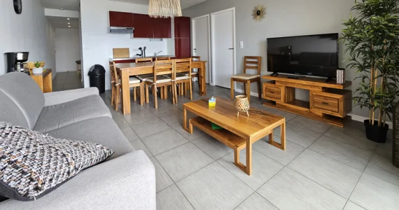 appart hôtel pasino n°3, pièce principale, au premier plan le coin salon et ses meuble en teck, son palmier et sa TV grand écran. Au fond une tables et ses chaises et la Kitchenette équipée aux façades rouges. A gauche une console en teck avec des éléments de décoration