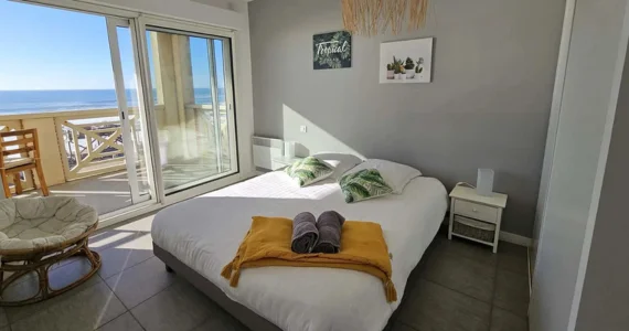 Appart hôtel Pasino n°4 à Lacanau Océan. Ici on découvre la chambre avec grande baie vitrée donnant directement sur la terrasse avec vue sur l'océan. Chambre moderne, décoration à dominante de gris perle composée d'un grand lit 2 places avec tables de chevets blanche composées d'un tiroir chacune.et d'un fauteuil en rotin. Sur le lit sont joliment déposés un dessus de lit plié jaune avec deux serviettes de bain grises roulées et posées dessus.