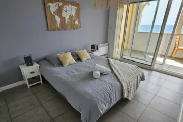 appart hôtel pasino n°3, chambre un grand lit 160X200 recouvert d'une couette grise et ses deux oreillers. Une chambre à coucher avec grand lit 160/200. Un plaid beige et deux serviettes de bain sont posés au coin du lit. La chambre ouvre sur la terrasse avec vue sur l'océan