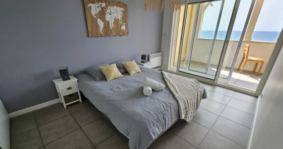appart hôtel pasino n°3, chambre un grand lit 160X200 recouvert d'une couette grise et ses deux oreillers. Une chambre à coucher avec grand lit 160/200. Un plaid beige et deux serviettes de bain sont posés au coin du lit. La chambre ouvre sur la terrasse avec vue sur l'océan