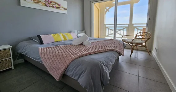 Appart hôtel Pasino n°6 chambre tons pastels, donnant sur la terrasse vue mer, 2 chevets, 2 lampes de chevet, 2 coussins décoration sur le lit, un plaid rose poudré et 2 serviettes de bain blanches, décorations au mur et plafond, un fauteuil en rotin et son coussin blanc. A l'arrière, la terrasse donnant sur l'océan