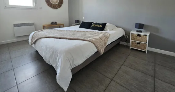 Appart hôtel Pasino n°6 Chambre N°3 murs blancs et gris, une décoration exotique sur le mur blanc dessous un banc en pin, une suspension au plafond, 2 chevets blancs : 2 tiroirs en Osiers plus lampes grises, un lit en 160X200 , 2 oreillers, 2 coussins décoration Vert foncé l'un marqué "Home", Un joli plaid beige est posé sur le lit