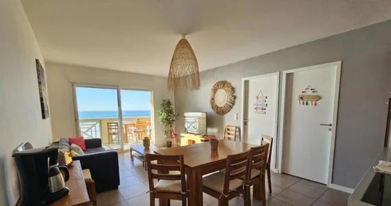 Appart hôtel Pasino n°4 Pièce à vivre, elle est lumineuse avec ses murs blancs. La pièce est meublée avec des meubles en teck: une table, ses six chaises et leurs coussins d'assise beiges, à gauche une console sur laquelle sont posés un grille-pain, une cafetière et une bouilloire. A l'arrière la partie salon : son canapé et ses coussins de décoration colorés, sa table de salon en teck , son meuble TV et la TV grand écran. Des éléments de décoration au mur, sur les 2 portes et au plafond, un palmier au fond de la pièce. une baie vitrée donne sur la terrasse vue mer et ses meubles en teck