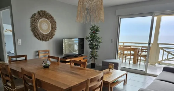 Appart hôtel Pasino n°6 pièce principale, au milieu une table en teck et ses 8 chaises, au fond le coin salon, une table, une TV grand écran, un pouf et un palmier décoration aux murs et plafond. A gauche un pouf et un palmier. La baie vitrée donne sur la grande terrasse équipée d'une table et de chaises vue mer.