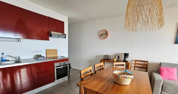 Appart hôtel Pasino N°1,pièce principale à gauche kitchenette rouge, au milieu une table en teck et ses chaises, au fond une console en teck sur laquelle sont posés une cafetière, un grille pain et une bouilloire, à droite un canapé et ses coussins. Au mur un tableau et une pendule