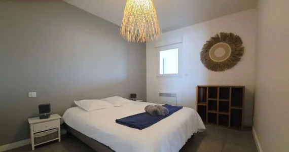 Appart hôtel Pasino n°5 Chambre. On y voit un grand lit 160/200 recouvert d'une couette avec housse blanche et ses 2 oreillers et leur taie. Un plaid bleu et 2 draps de douches gris sont posés sur la couette. Deux chevets à tiroir attenants au lit. Côté mur, une fenêtre donnant sur le palier et un meuble de rangement en teck. Décoration exotique