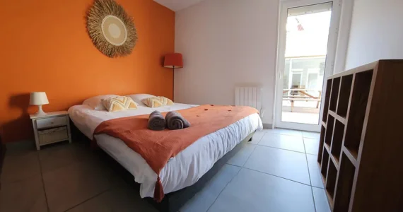 Appart hôtel Pasino n°7 chambre avec grand lit double 160x200 donnant sur la patio. Décoration dans les tons orangés avec plaid orange sur le lit et 2 serviettes de bain grise. Chambre entièrement carrelée au sol (couleur grise)